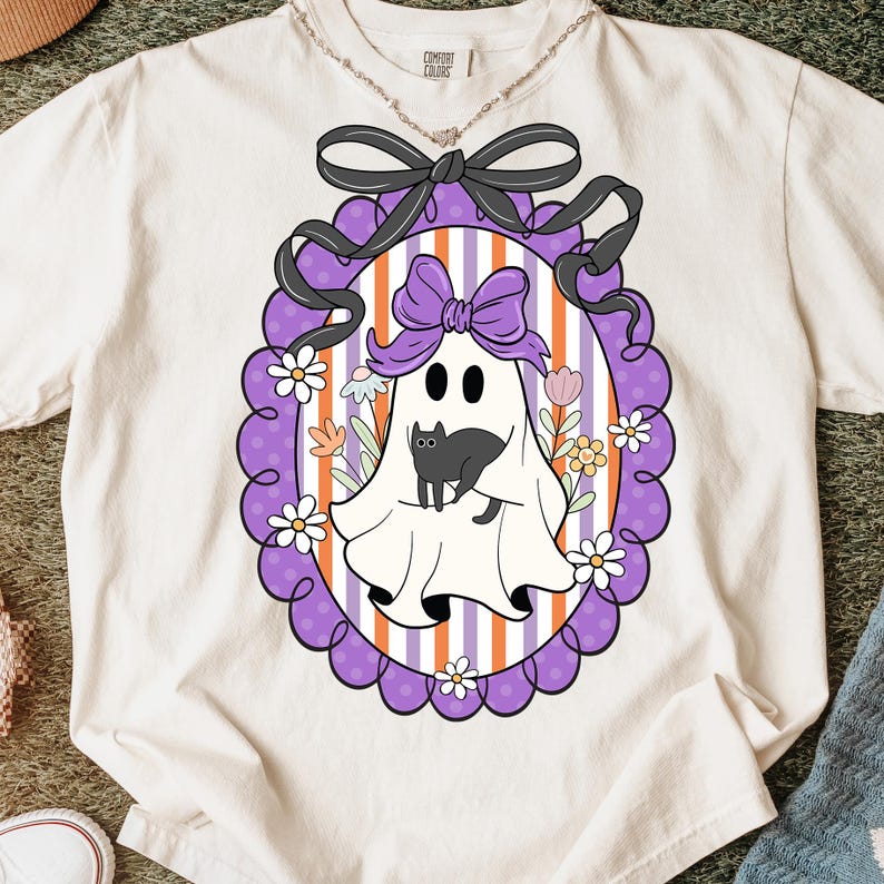 May include: Cream-colored t-shirt featuring a Halloween-themed graphic. The graphic includes a white ghost with a black cat, a purple bow, and a decorative frame with flowers and stripes. The shirt has a Comfort Colors label.