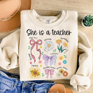 Op de afbeelding: Cr&egrave;mekleurige sweatshirt met de tekst "She is a teacher" en bloemen- en strik-afbeeldingen. Het ontwerp bevat de woorden "Chosen", "Beautiful", "Victorious", "Capable", "Greatly Loved" en "Never Alone".