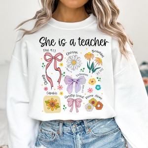 Op de afbeelding: Witte sweatshirt met de tekst "She is a teacher" in het zwart. Het ontwerp bevat bloemen- en strik-afbeeldingen met woorden als "Chosen", "Beautiful" en "Victorious".