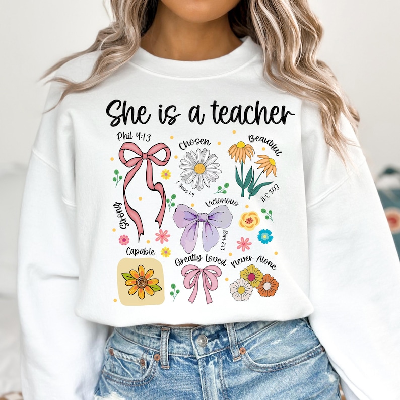 Op de afbeelding: Witte sweatshirt met de tekst "She is a teacher" in het zwart. Het ontwerp bevat bloemen- en strik-afbeeldingen met woorden als "Chosen", "Beautiful" en "Victorious".