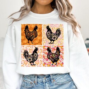 May include: White sweatshirt featuring a four-panel design with black rooster silhouettes filled with floral patterns. The background panels showcase pumpkins in orange and pink hues, creating a fall-themed aesthetic.