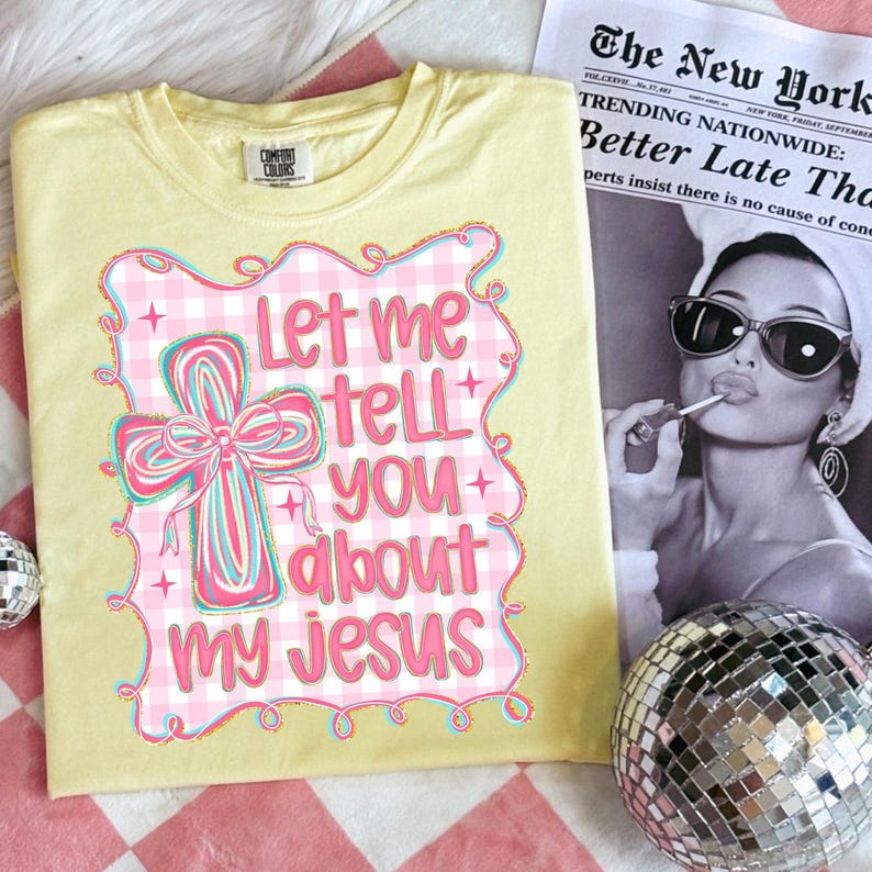 May include: A light yellow Comfort Colors t-shirt with a graphic that says "Let me tell you about my Jesus." The graphic features a cross with a pink bow and a pink and white gingham border. A newspaper and disco ball are in the background.