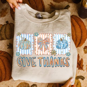 May include: A beige sweatshirt featuring a colorful graphic with pumpkins, a bow, and the text "GIVE THANKS" in orange and blue. The design includes blue and orange accents, perfect for fall.