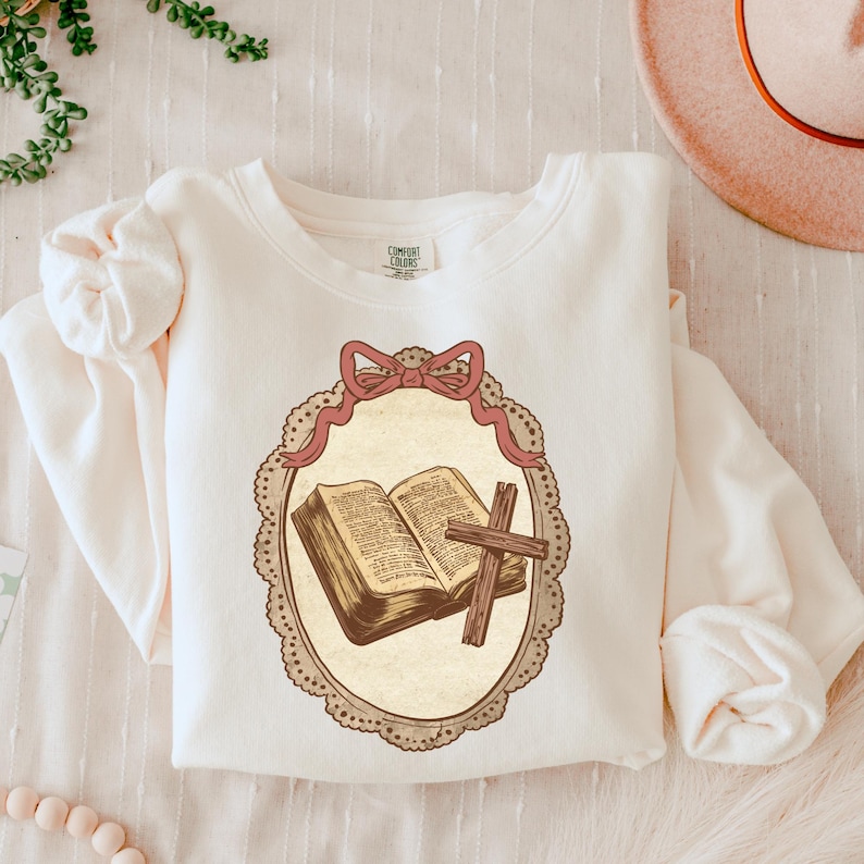 May include: Cream-colored sweatshirt featuring a framed illustration of an open book and a wooden cross. The illustration is in a vintage style, with a pink bow at the top of the frame. The sweatshirt has long sleeves and a crew neck.