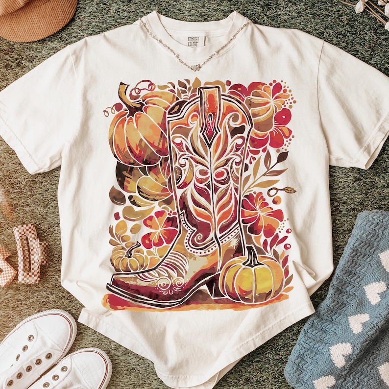 Op de afbeelding: Cr&egrave;mekleurig T-shirt met een kleurrijke afbeelding van een cowboylaars, pompoenen en bloemelementen in tinten oranje, bruin en rood. Het shirt heeft een relaxte pasvorm en korte mouwen. De tekst "COMFORT COLORS" is zichtbaar op de kraag.