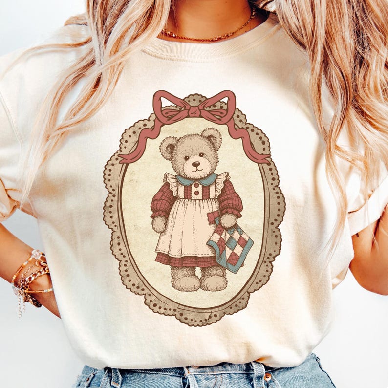 Op de afbeelding: Cr&egrave;mekleurig T-shirt met een vintage-stijl illustratie van een teddybeer in een ovale lijst. De beer draagt een jurk en houdt een quilt vast. De lijst heeft een decoratieve strik aan de bovenkant.
