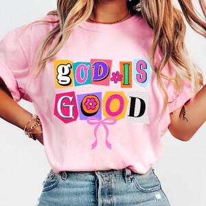 May include: A light pink t-shirt with the words "GOD IS GOOD" in a colorful, cut-out style design. The letters are in various colors, with a flower and a pink bow. The shirt is made of a soft material.