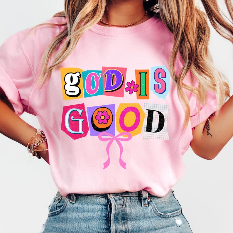 May include: A light pink t-shirt with the words "GOD IS GOOD" in a colorful, cut-out style design. The letters are in various colors, with a flower and a pink bow. The shirt is made of a soft material.