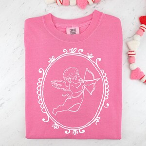 Pu&ograve; includere: Una felpa rosa con una grafica bianca di Cupido all'interno di una cornice ovale. La felpa ha la scritta "Comfort Colors" sull'etichetta. Il design ha un tema romantico, perfetto per San Valentino o come regalo.