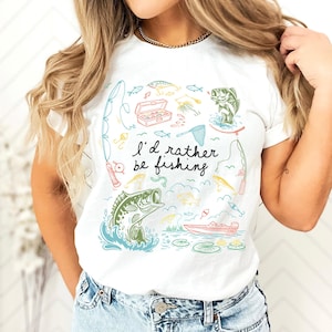 May include: White t-shirt with a colorful fishing-themed graphic. The design includes illustrations of fish, fishing rods, a tackle box, and a boat. The text "I'd rather be fishing" is also featured.