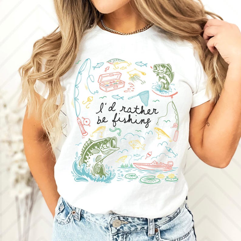 May include: White t-shirt with a colorful fishing-themed graphic. The design includes illustrations of fish, fishing rods, a tackle box, and a boat. The text "I'd rather be fishing" is also featured.