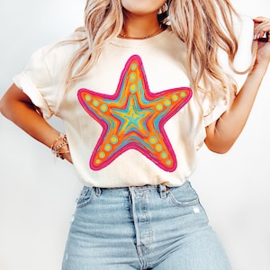 May include: Cream-colored t-shirt featuring a vibrant, colorful starfish design. The starfish has a pink outline, with orange, yellow, and turquoise details. The shirt is paired with blue jeans.