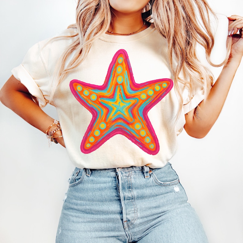 May include: Cream-colored t-shirt featuring a vibrant, colorful starfish design. The starfish has a pink outline, with orange, yellow, and turquoise details. The shirt is paired with blue jeans.