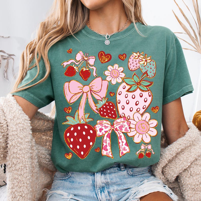 Op de afbeelding: Een groen T-shirt met een grafische print van aardbeien, strikken en bloemen. De aardbeien zijn rood met witte zaadjes en de strikken zijn roze met gouden accenten.