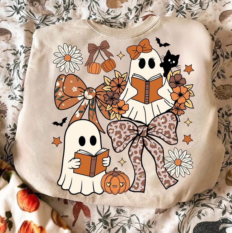 Op de afbeelding: Beige sweatshirt met een Halloween-thema. Voorzien van twee stripfiguurgeesten die boeken lezen, omringd door strikken, pompoenen, bloemen, sterren en een zwarte kat. Het ontwerp bevat oranje, bruine en witte kleuren.