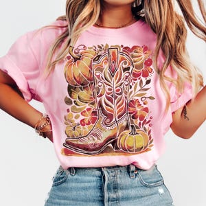 Op de afbeelding: Roze t-shirt met een herfstthema. Het ontwerp toont een gedetailleerde cowboylaars, pompoenen en bloemelementen in bruin, oranje en rode tinten. Het shirt is gemaakt van een zacht materiaal en heeft korte mouwen.