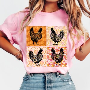 May include: Pink t-shirt featuring a square design with four black rooster illustrations. Each rooster is filled with floral patterns and set against a background of pumpkins and flowers. The shirt is a light pink color.