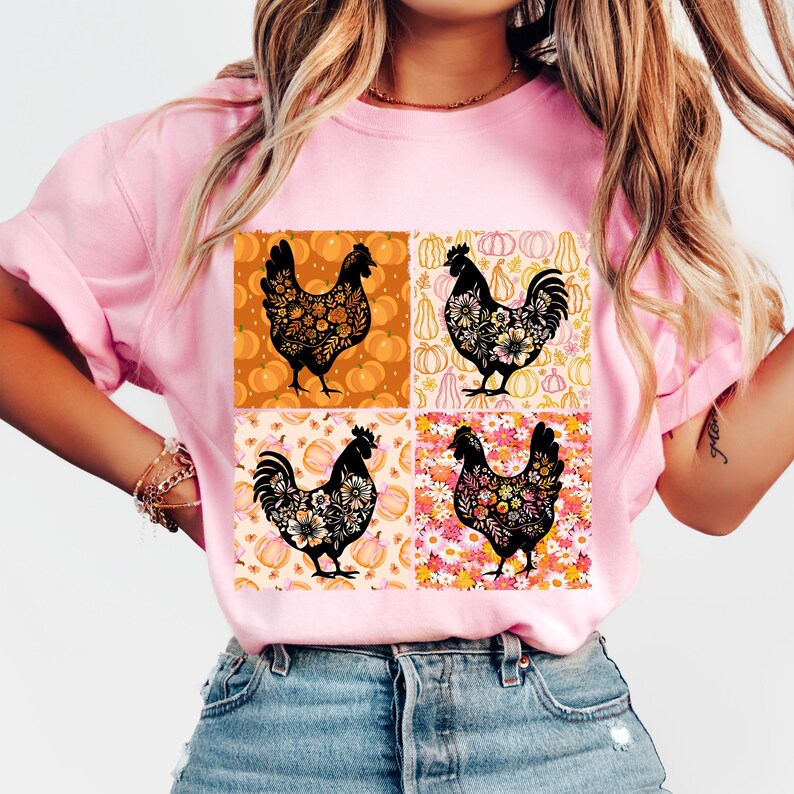 May include: Pink t-shirt featuring a square design with four black rooster illustrations. Each rooster is filled with floral patterns and set against a background of pumpkins and flowers. The shirt is a light pink color.