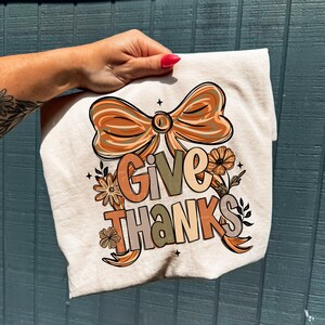 May include: A cream-colored t-shirt featuring the words "GIVE THANKS" in a fall-themed design. The design includes a large bow, flowers, and leaves in shades of orange, brown, and green. The shirt is held by a hand with a red nail polish.