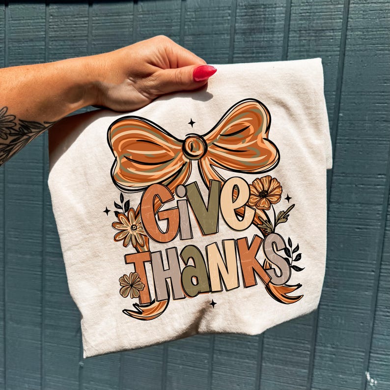 May include: A cream-colored t-shirt featuring the words "GIVE THANKS" in a fall-themed design. The design includes a large bow, flowers, and leaves in shades of orange, brown, and green. The shirt is held by a hand with a red nail polish.