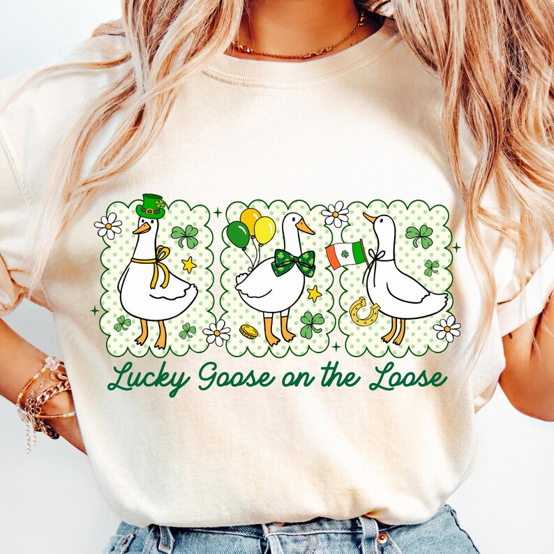 Peut inclure: T-shirt cr&egrave;me avec trois oies de dessin anim&eacute;, chacune avec des accessoires de la Saint-Patrick. Les oies sont dans une bordure verte festonn&eacute;e avec des tr&egrave;fles, des fleurs et des &eacute;toiles. Le texte dit "Lucky Goose on the Loose" en vert.