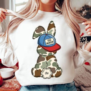 May include: White sweatshirt with a cartoon bunny wearing a blue and red baseball cap that says "Egg Hunter". The bunny is camouflage.
