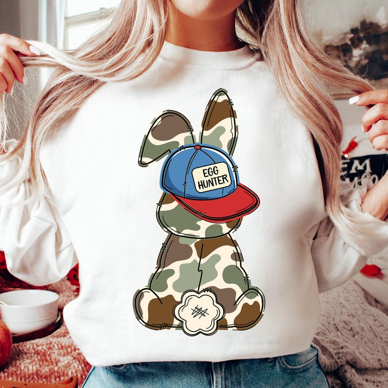 May include: White sweatshirt with a cartoon bunny wearing a blue and red baseball cap that says "Egg Hunter". The bunny is camouflage.