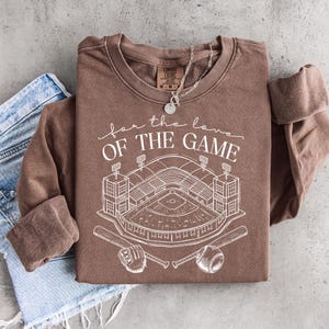 Op de afbeelding: Een bruine sweatshirt met lange mouwen met de tekst "for the love of the game" en een witte honkbalstadionafbeelding. Het ontwerp omvat honkbalknuppels, een honkbal en een handschoen. Een zilveren ketting is over de trui gedrapeerd, die op een spijkerbroek is gevouwen.