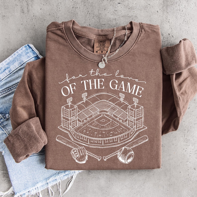 Op de afbeelding: Een bruine sweatshirt met lange mouwen met de tekst "for the love of the game" en een witte honkbalstadionafbeelding. Het ontwerp omvat honkbalknuppels, een honkbal en een handschoen. Een zilveren ketting is over de trui gedrapeerd, die op een spijkerbroek is gevouwen.