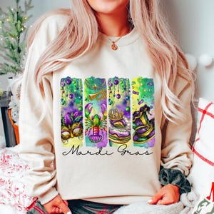 May include: A cream-colored sweatshirt with a colorful Mardi Gras design. The design features a crown, a trumpet, a lobster, a mask, and a hat. The text "Mardi Gras" is printed below the design.