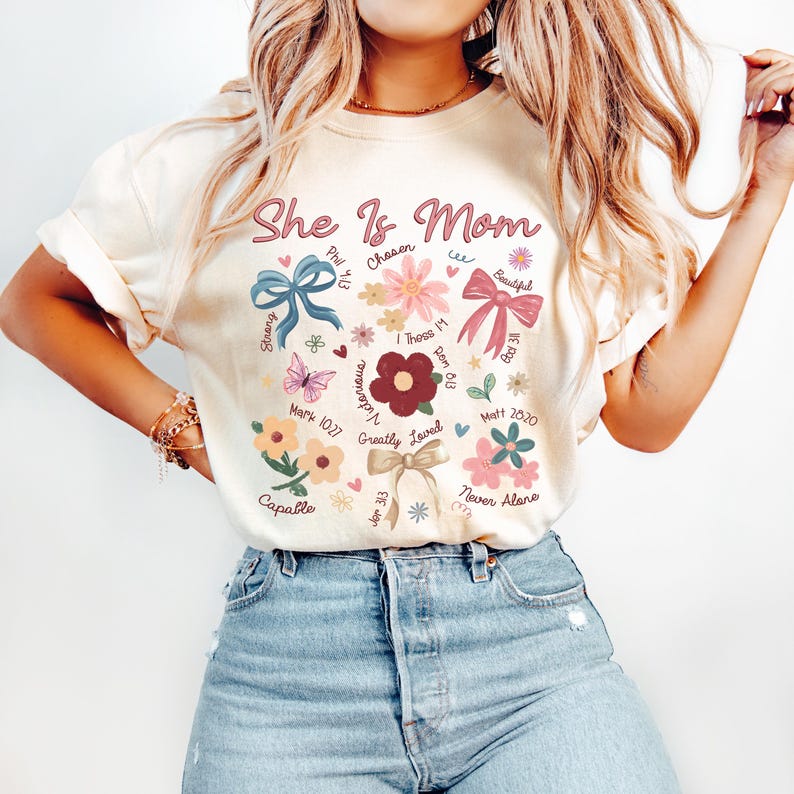 K&ouml;nnte beinhalten: Ein cremefarbenes T-Shirt mit dem Text "She Is Mom" in rosa und blauen Buchstaben. Das Shirt zeigt ein Blumenmuster mit verschiedenen Bibelversen und W&ouml;rtern wie "Chosen", "Strong", "Victorious", "Greatly Loved", "Capable", "Never Alone" und "Beautiful".
