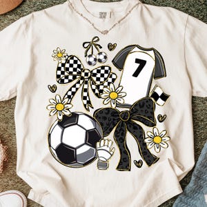 May include: Cream-colored t-shirt with a soccer-themed design. Features a soccer ball, jersey with the number 7, bows, flowers, and hearts. The design incorporates black, white, and gold accents, creating a playful and sporty aesthetic.