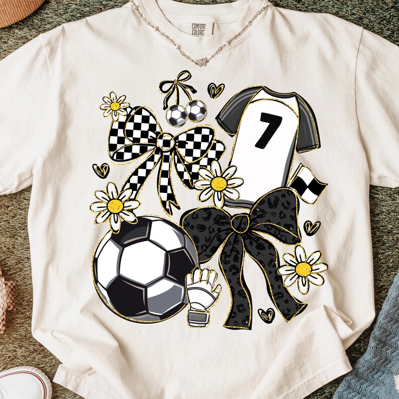 May include: Cream-colored t-shirt with a soccer-themed design. Features a soccer ball, jersey with the number 7, bows, flowers, and hearts. The design incorporates black, white, and gold accents, creating a playful and sporty aesthetic.