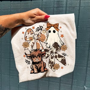 Op de afbeelding: Beige grafisch T-shirt met een cartoon spook, een Schotse Hooglandkoe en twee grote strikken met bloemen- en koeienprintpatronen. Het ontwerp bevat sterren en bloemen in bruin-, oranje- en cr&egrave;metinten.