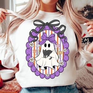 May include: White sweatshirt featuring a Halloween-themed graphic. The design includes a cartoon ghost with a black cat, surrounded by flowers, inside a purple frame with a black bow. The background has purple and orange stripes.