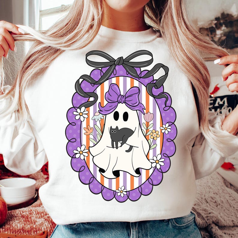 May include: White sweatshirt featuring a Halloween-themed graphic. The design includes a cartoon ghost with a black cat, surrounded by flowers, inside a purple frame with a black bow. The background has purple and orange stripes.