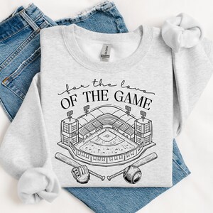 Op de afbeelding: Een lichtgrijs sweatshirt met de tekst "for the love of the game" boven een honkbalstadionafbeelding. De afbeelding bevat een honkbal, een knuppel en een handschoen. Het sweatshirt is op een blauwe jeans gevouwen.