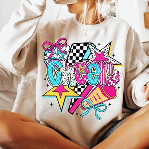 Op de afbeelding: Een beige sweatshirt met een kleurrijke "Cheer"-afbeelding. Het ontwerp bevat sterren, een megafoon en een strik. Het woord "Cheer" is in verschillende lettertypen en kleuren, waaronder blauw, roze en geel.