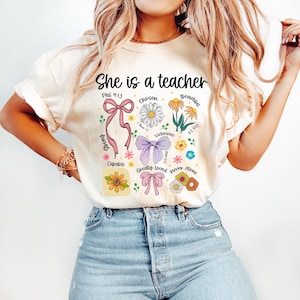 Op de afbeelding: Cr&egrave;mekleurig T-shirt met de tekst "She is a teacher" en bloemen- en lintafbeeldingen. Het ontwerp bevat de woorden "Chosen", "Victorious", "Beautiful" en "Greatly Loved".