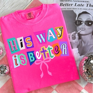 May include: A bright pink t-shirt with the phrase "His Way is Better" in colorful, cut-out style letters. A pink bow is below the text. The shirt is on a pink and white checkered surface.