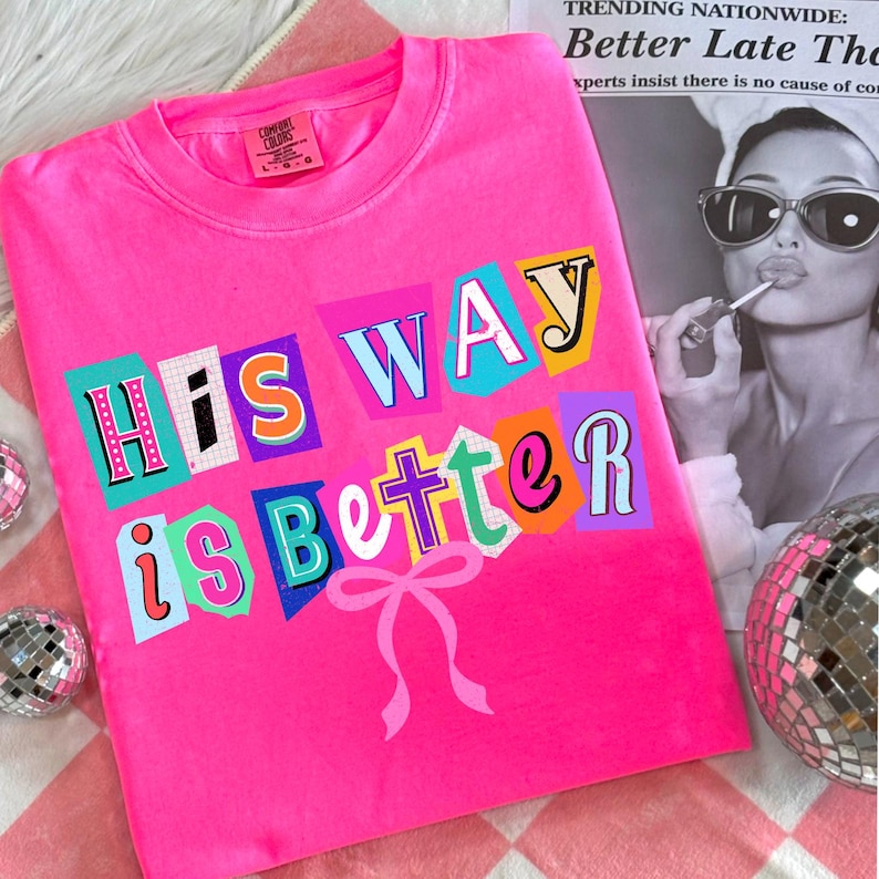 May include: A bright pink t-shirt with the phrase "His Way is Better" in colorful, cut-out style letters. A pink bow is below the text. The shirt is on a pink and white checkered surface.