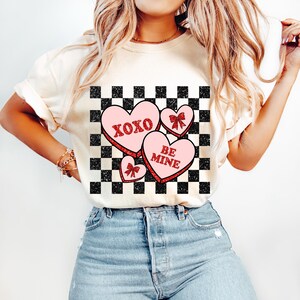 May include: White t-shirt with a checkered pattern and three pink hearts with red glitter. The hearts have the text "XOXO" and "BE MINE" on them.