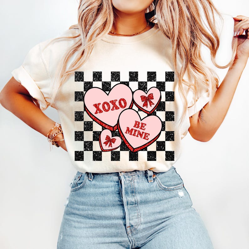 May include: White t-shirt with a checkered pattern and three pink hearts with red glitter. The hearts have the text "XOXO" and "BE MINE" on them.
