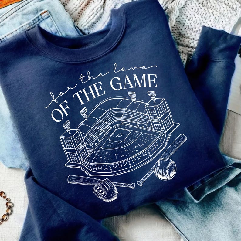 Op de afbeelding: Marineblauw sweatshirt met een witte afbeelding van een honkbalstadion, honkbal, handschoen en knuppels. De tekst "for the love of the game" staat boven het stadionontwerp. Het sweatshirt is op een blauwe spijkerbroek gevouwen.