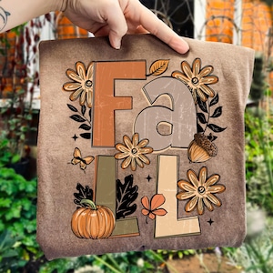May include: A brown t-shirt featuring the word "FALL" in a decorative font with fall-themed illustrations. The design includes flowers, a pumpkin, an acorn, a butterfly, and leaves in shades of orange, brown, and green.