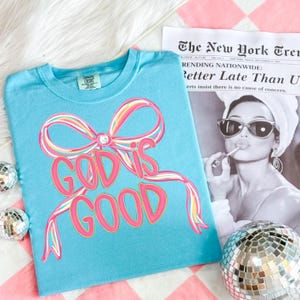 May include: A light blue t-shirt with a pink and yellow bow design and the words "GOD IS GOOD" in pink. The shirt is folded on a pink and white checkered surface, with a newspaper and disco ball nearby.