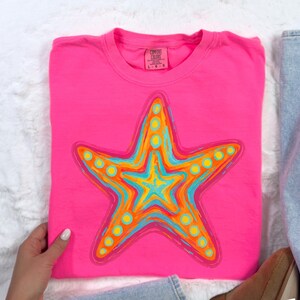 May include: A hot pink t-shirt featuring a colorful starfish design. The starfish is outlined in dark blue and filled with shades of orange, yellow, and turquoise. The shirt is folded and has a Comfort Colors tag.