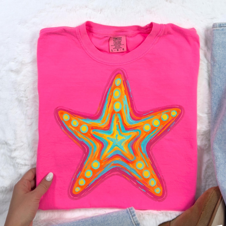 May include: A hot pink t-shirt featuring a colorful starfish design. The starfish is outlined in dark blue and filled with shades of orange, yellow, and turquoise. The shirt is folded and has a Comfort Colors tag.