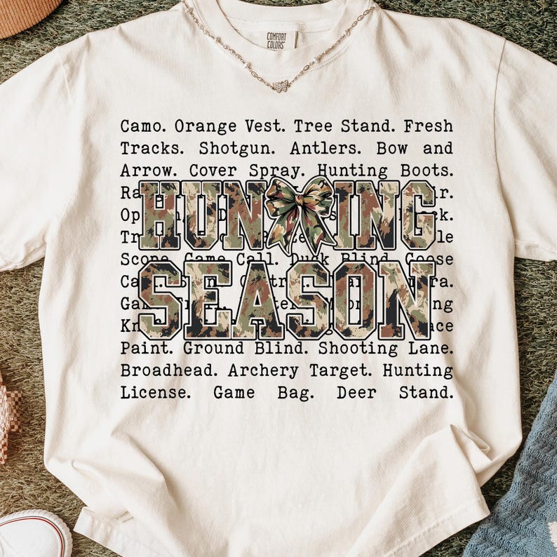 May include: Cream-colored t-shirt with the words "HUNTING SEASON" in a camouflage design. The shirt also lists hunting-related terms like "Shotgun," "Antlers," and "Bow and Arrow."