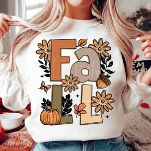 May include: White sweatshirt with the word "FALL" in large, distressed letters. The design includes fall-themed graphics like flowers, a pumpkin, a butterfly, and an acorn. The letters are in shades of brown, green, and beige.